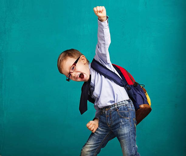 Cheerful smiling little boy with big backpack jumping and having fun against blue wall. Looking at camera. School concept. Back to School